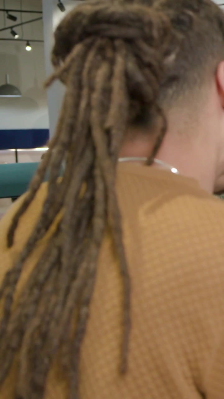 Video verticale: Telecamera che segue un uomo che cammina attraverso un salone, mostrando i suoi dreadlocks e un top marrone senape