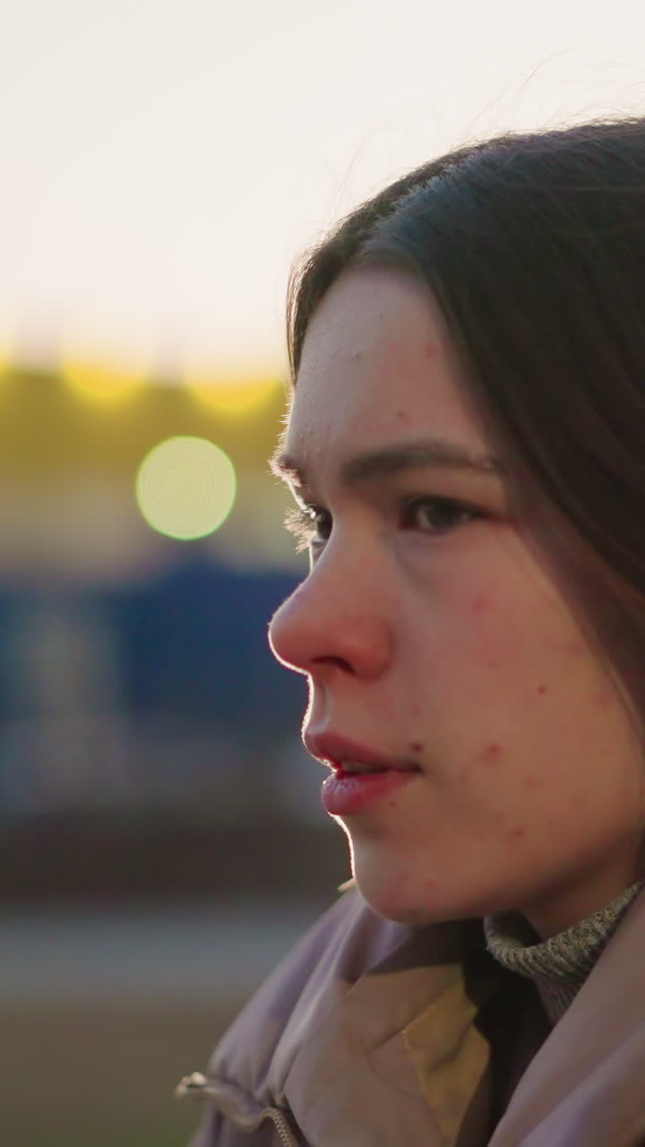 fotografía lateral de una mujer joven con una chaqueta de melocotón, con la boca ligeramente abierta, perdida en sus pensamientos. la suave luz de la tarde y el fondo urbano borroso se suman al estado de ánimo contemplativo