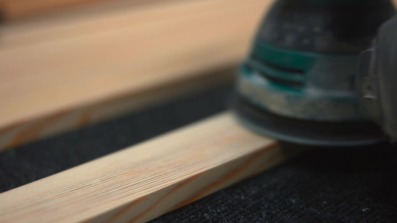 Primo piano di una levigatrice orbitale manuale a diretto contatto con un listello di pino chiaro su un supporto scuro, che mostra il platorello circolare, polvere sottile, il bordo del guanto dell'operatore, texture usurate, riflessi caldi, sfocatura morbida e