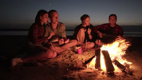 Friends-company-of-four-are-hanging-out-on-beach-near-the-bonfire.-taking-sausages,-drinking,-playing-the-guitar.-Dark-sea