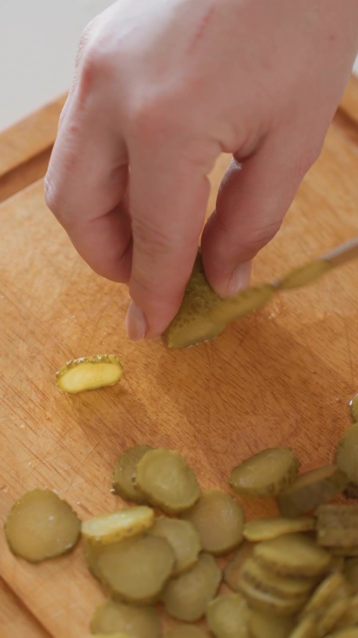 Primo piano obliquo dall'alto di una mano che affetta cetriolini in fette sottili e rotonde su un tagliere di legno caldo, mostrando la preparazione fresca in cucina, la consistenza verde e umida, il controllo del coltello piccolo e il movimento attento.