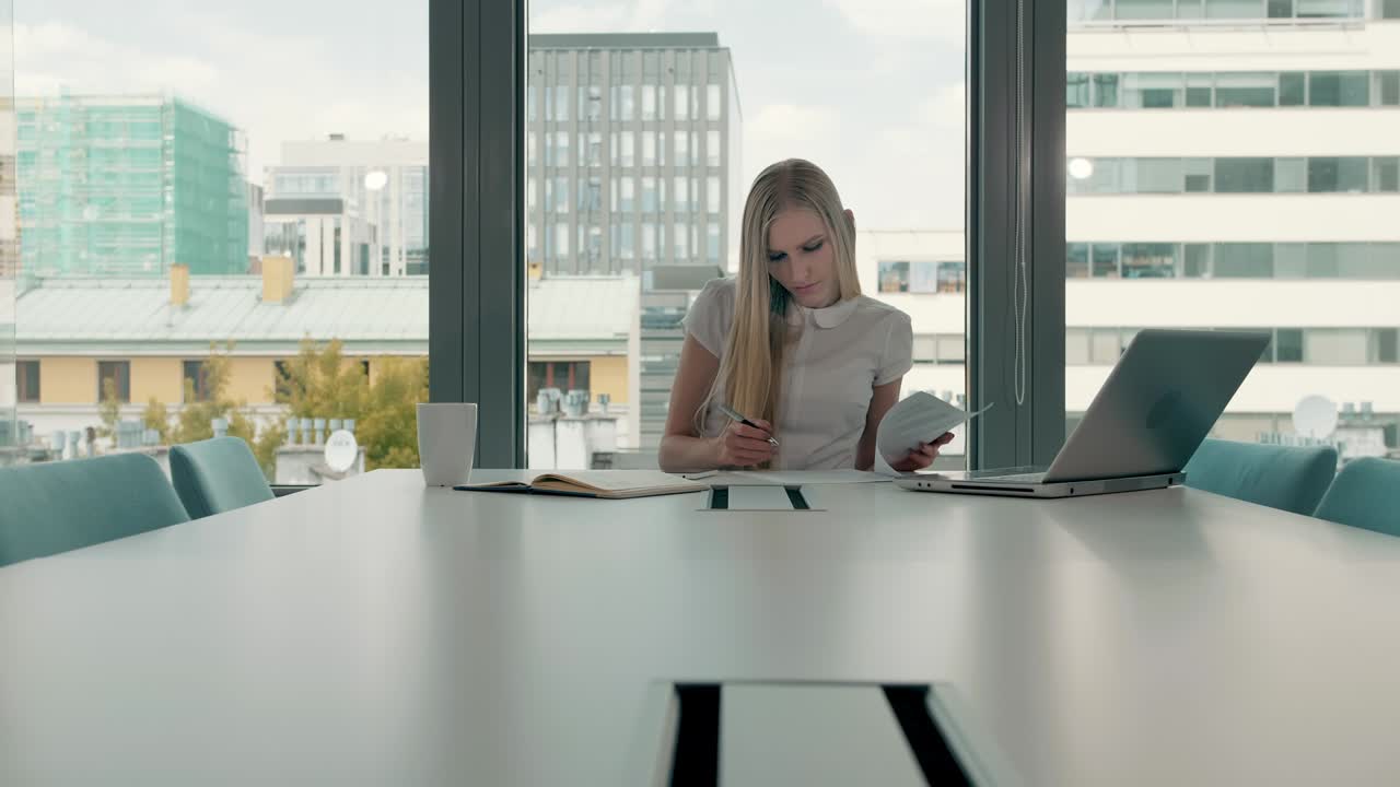 Młoda bizneswoman w sali konferencyjnej z dokumentami. Elegancka blondynka siedząca przy długim stole w nowoczesnej sali biurowej z laptopem i pisząca na papierach podczas pracy.