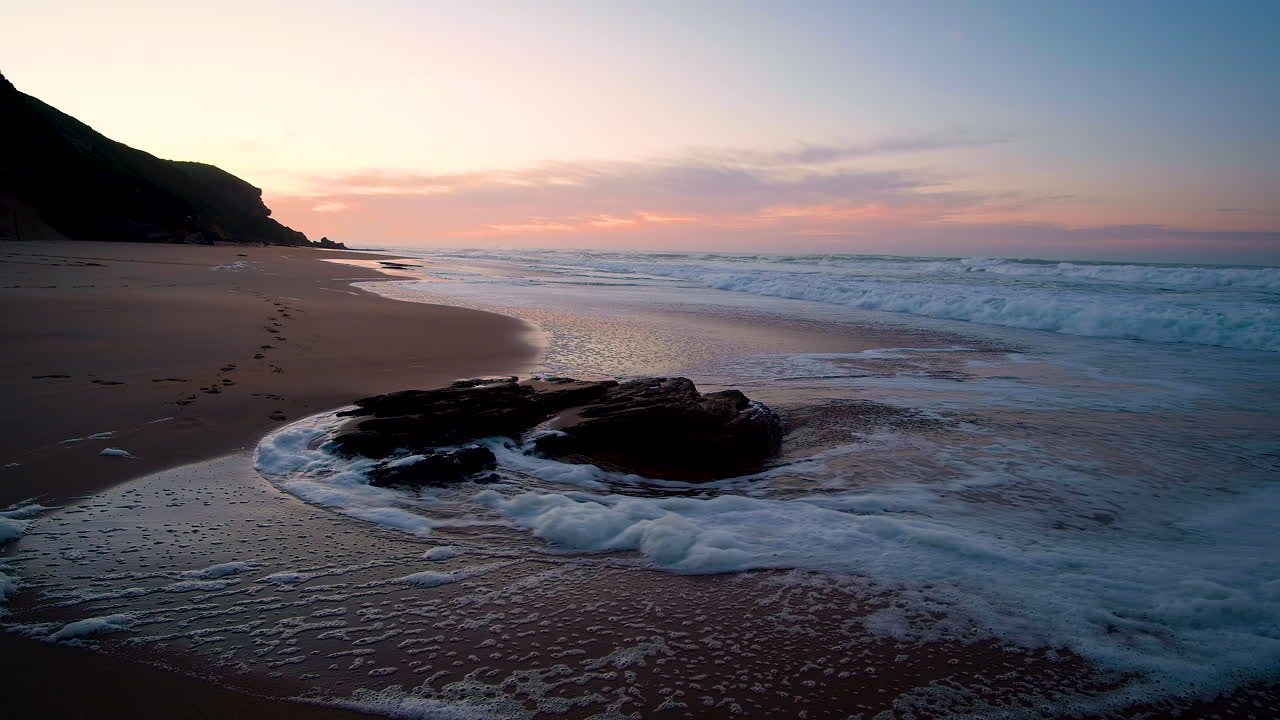Small wave gently envelops lone rock on beach during vibrant sunrise