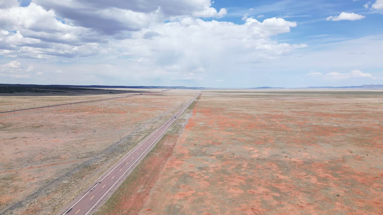 Drone pans to follow minivan down route 66 as it slowly rises and reveals the incredibly vast and flat plain. There is a single two-lane road with puffy clouds in a blue sky on a sunny day