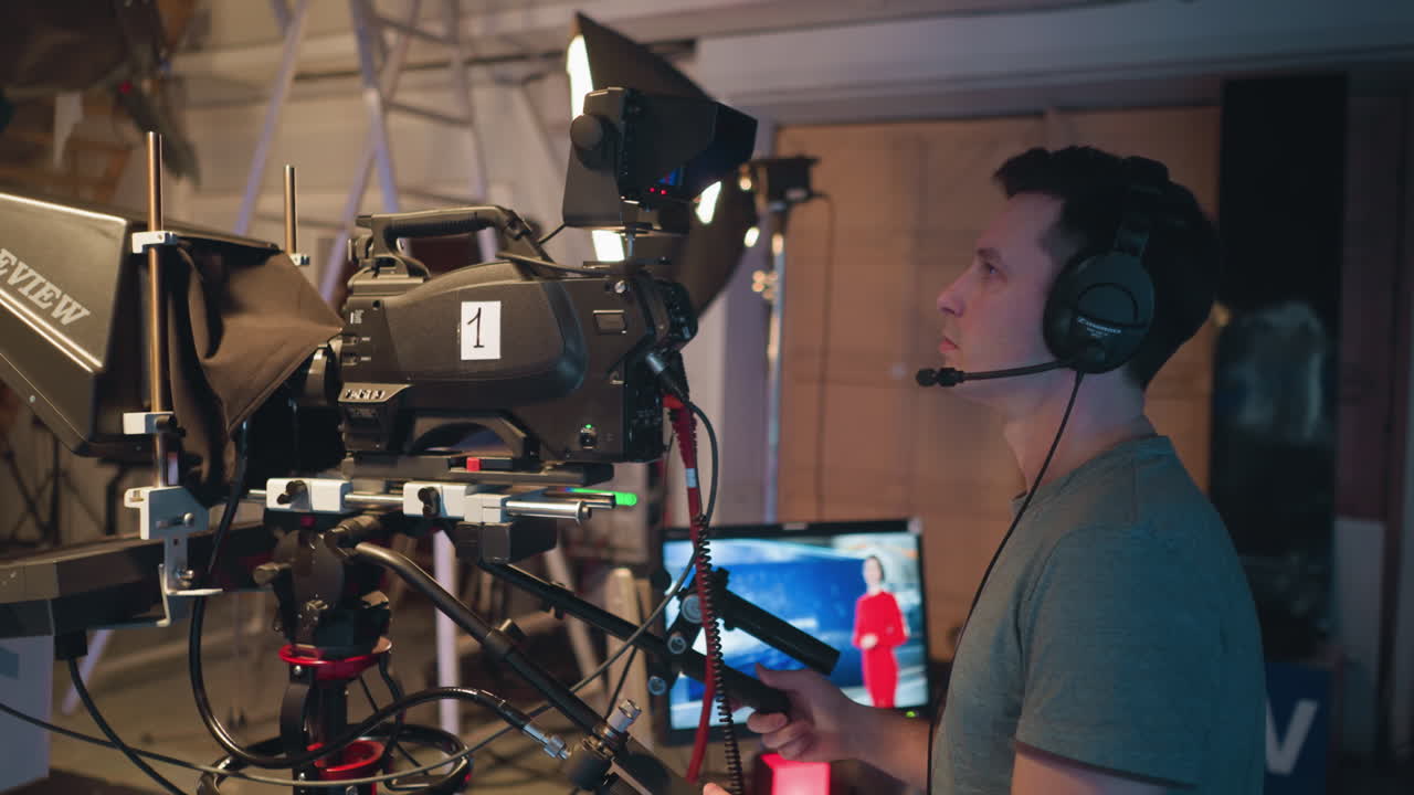 Male cameraman wearing headset operates professional broadcast camera in television studio during video production with teleprompter and soft lighting equipment while monitoring presenter performance on screen