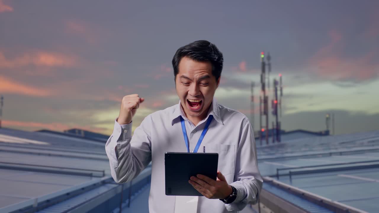 工業用屋上ソーラーパネルを点検しながらタブレットを手に立つアジア人男性専門作業員が、目標達成の雄叫びを上げて拳を突き上げている。