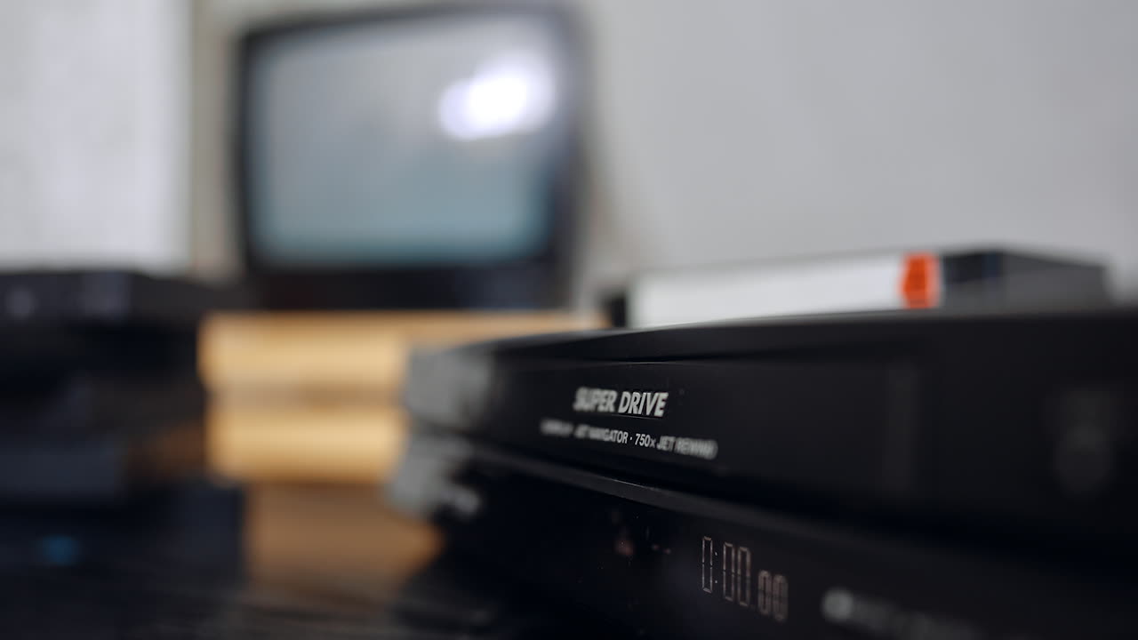 Una cassetta VHS viene inserita nel lettore. Primo piano. Lo schermo sullo sfondo sfocato inizia a mostrare l'immagine.
