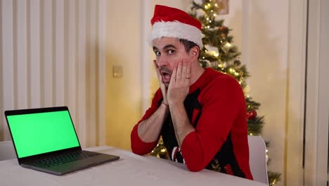 festive male wearing christmas hat and sweater looking at camera and laptop computer green screen with surprised shock expressions not believing what he has seen