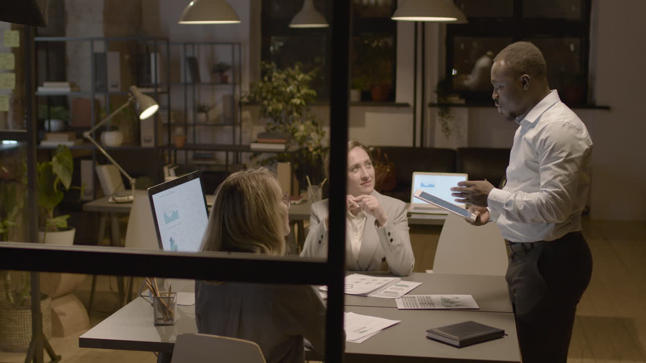 American Man Employee Holding A Tablet And Explaining A Project To Female Coworkers Who Are Sitting At Desk In The Office