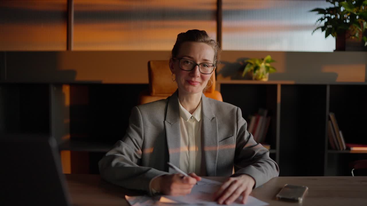 Portrait of a confident middle-aged blonde businesswoman with glasses in a gray jacket who sits in front of sheets of papers with a writing pen in her hands at a wooden table in a sunny office