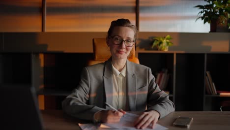 Portrait-of-a-confident-middle-aged-blonde-businesswoman-with-glasses-in-a-gray-jacket-who-sits-in-front-of-sheets-of-papers-with-a-writing-pen-in-her-hands-at-a-wooden-table-in-a-sunny-office