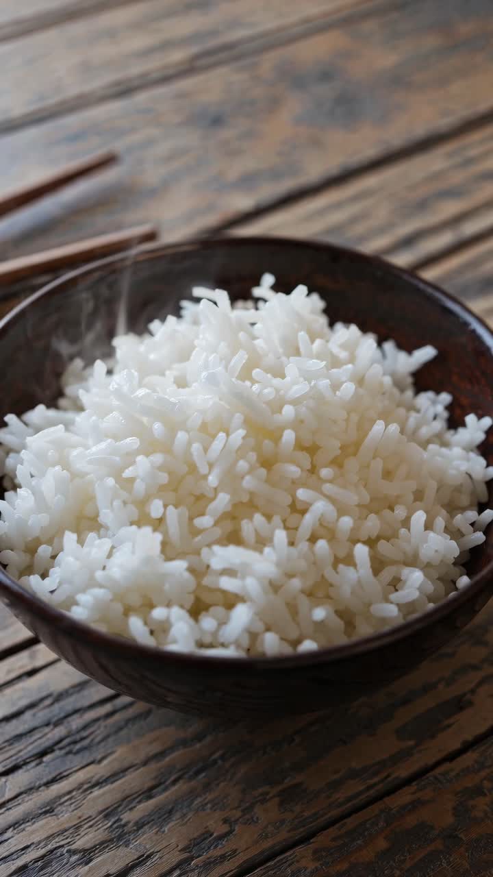 Close-up video shot of steaming white rice in a dark bowl on a rustic wooden table