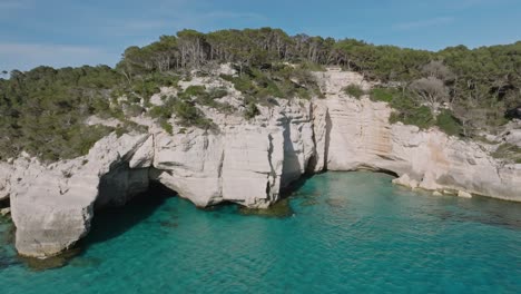 hidden trail leads down to mitjana beach in menorca spain where perfect white limestone cliffs can be seen and luscious forest surrounding