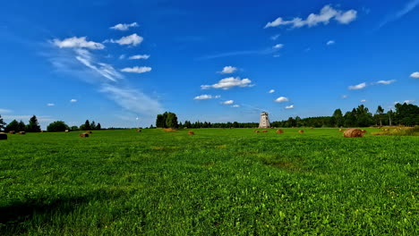 Camine-Por-Una-Relajante-Pradera-Cubierta-De-Sol,-Con-Un-Antiguo-Molino-De-Viento-Letón-Al-Fondo