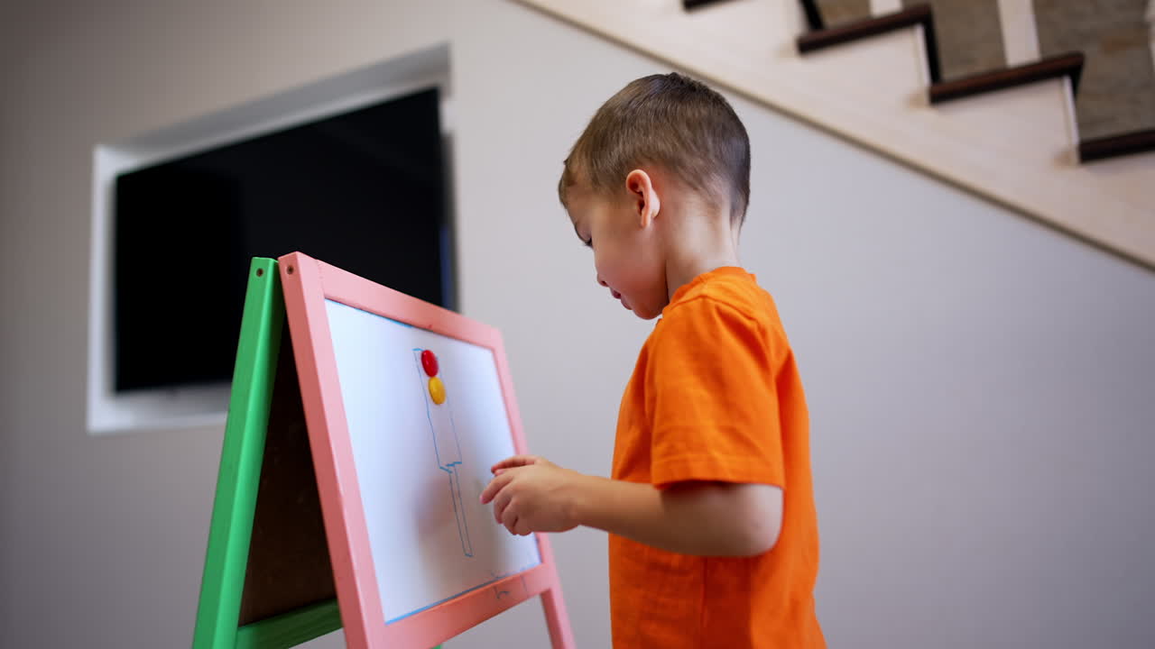 Little toddler boy in orange t-shirt stands at whiteboard. Kid draws a traffic light and puts colorful magnets on.