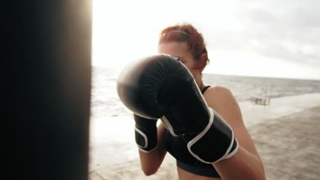 close up view of a strong athletic female boxer in gloves exercising with a bag against the son by the sea. female boxer training