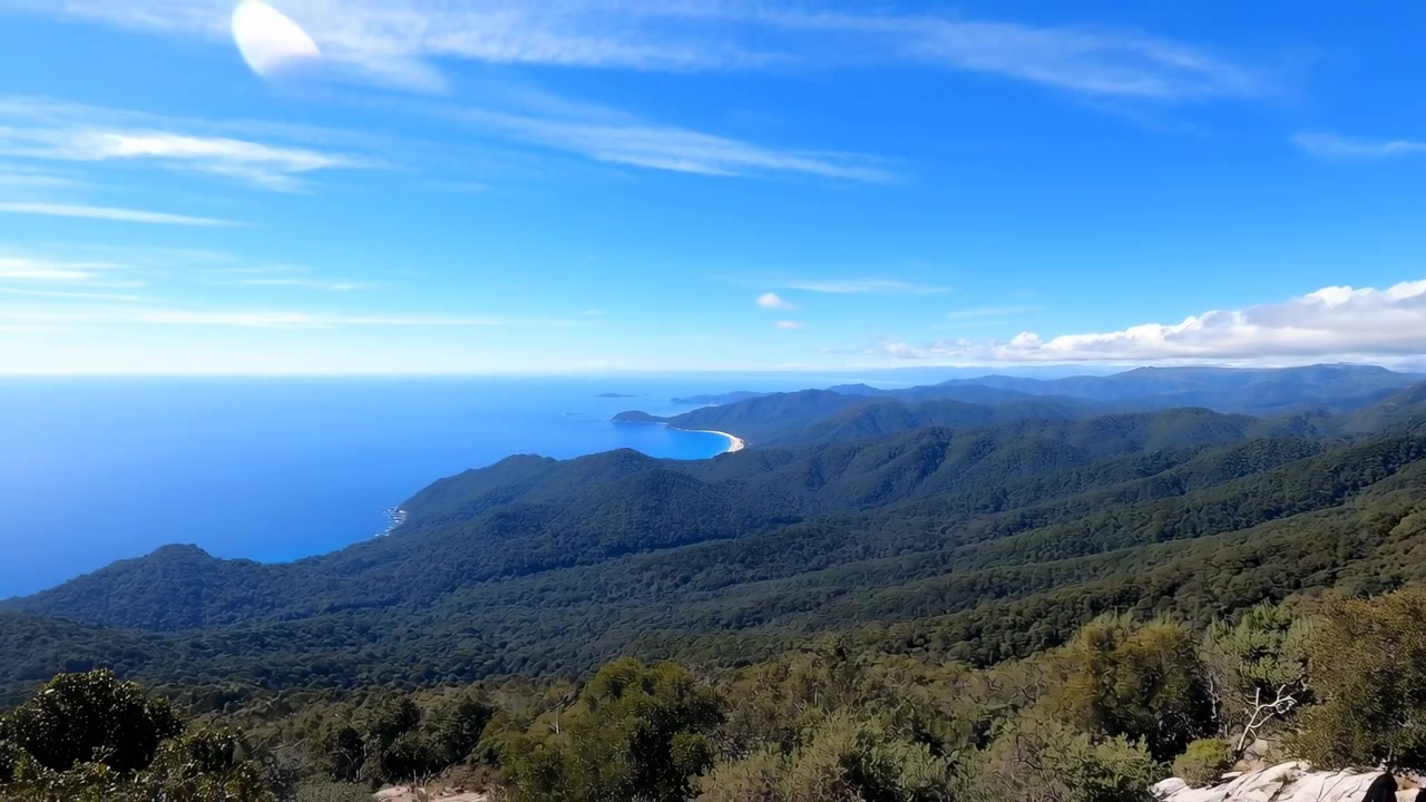 Una impresionante vista panorámica de una cordillera con un cielo azul claro y el vasto océano de fondo. Este sereno y expansivo paisaje natural presenta terreno rocoso y exuberante vegetación.