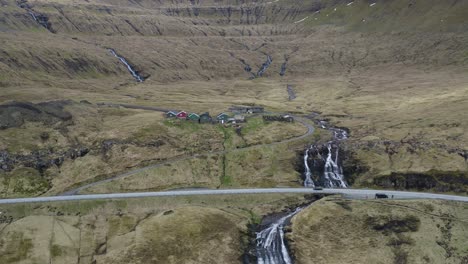 Vista-Aérea-De-La-Idílica-Cascada-Que-Fluye-Cuesta-Abajo-En-Fuglafjörður-En-La-Isla-De-Eysturoy,-Islas-Feroe---Espectacular-Toma-De-Drones-Con-Autos-En-Carretera-De-Montaña