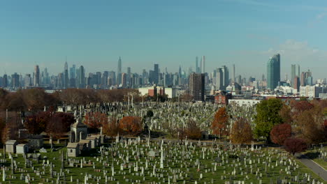 Forwards-fly-above-historic-Calvary-Cemetery.-Old-tombstones-on-green-lawn-between-autumn-coloured-trees.-Skyline-with-Manhattan-skyscrapers.-Queens,-New-York-City,-USA