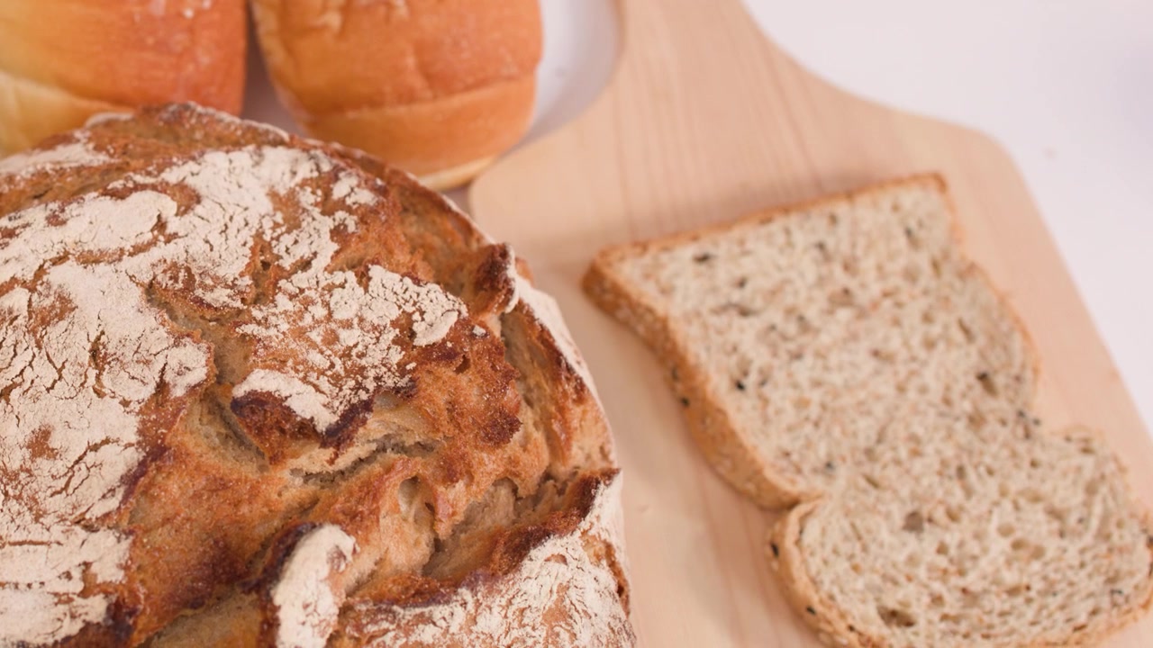 A hand places a multigrain bread slice onto a wooden board beside fresh bakery assortments.