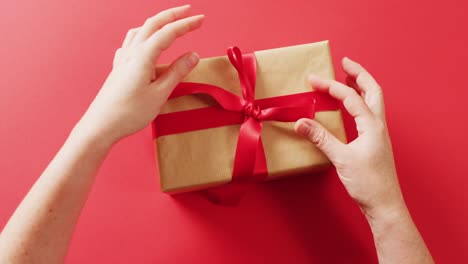 Video-of-hands-tying-red-ribbon-on-gift-box-wrapped-in-brown-paper,-on-red-background