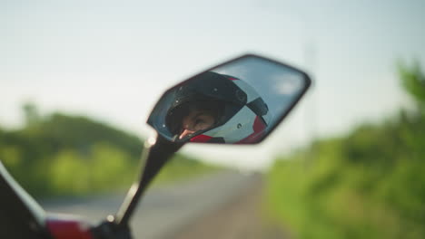primer plano de la cara de un motociclista visto a través de un espejo de motocicleta, con un casco, el fondo presenta un borrón suave de vegetación exuberante y una carretera