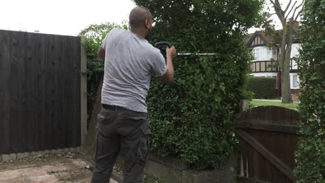 uk asian adult bald male using electric hedge trimmer outside in front garden