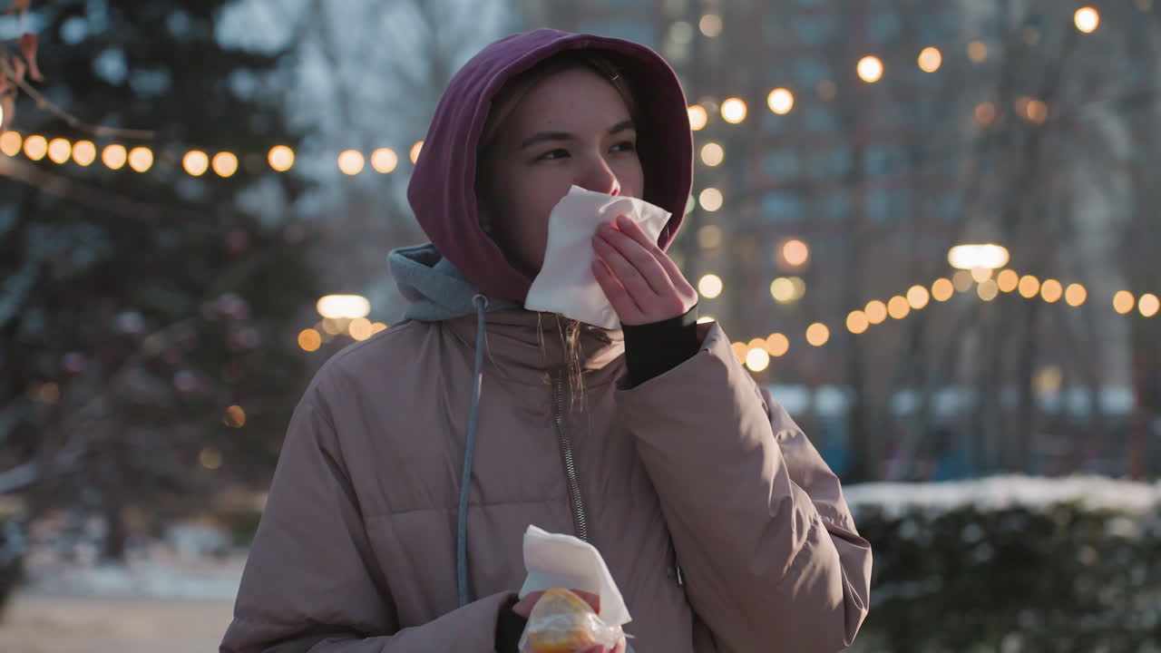 손에 킨을 들고 간식을 들고 있는 여성, 배경에 보케 조명과 함께 야외에서 입을 청소하고, 추운 겨울철, 거리 음식을 즐기고, 맛있는 간식을 먹은 후 입을 고 있습니다.