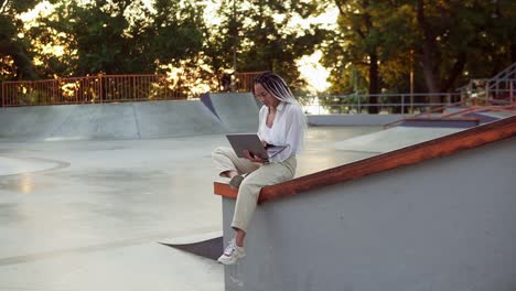 Young-elegant-caucasian-woman-with-dreadlocks-in-white-blouse-working-in-summer-park.-Female-freelancer-typing-on-laptop