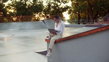 young elegant caucasian woman with dreadlocks in white blouse working in summer park. female freelancer typing on laptop