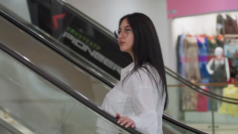 side view of woman wearing white shirt and glasses stepping onto ascending escalator with hand placed on railing, surrounded by colorful clothing displays and mannequins inside vibrant shopping mall