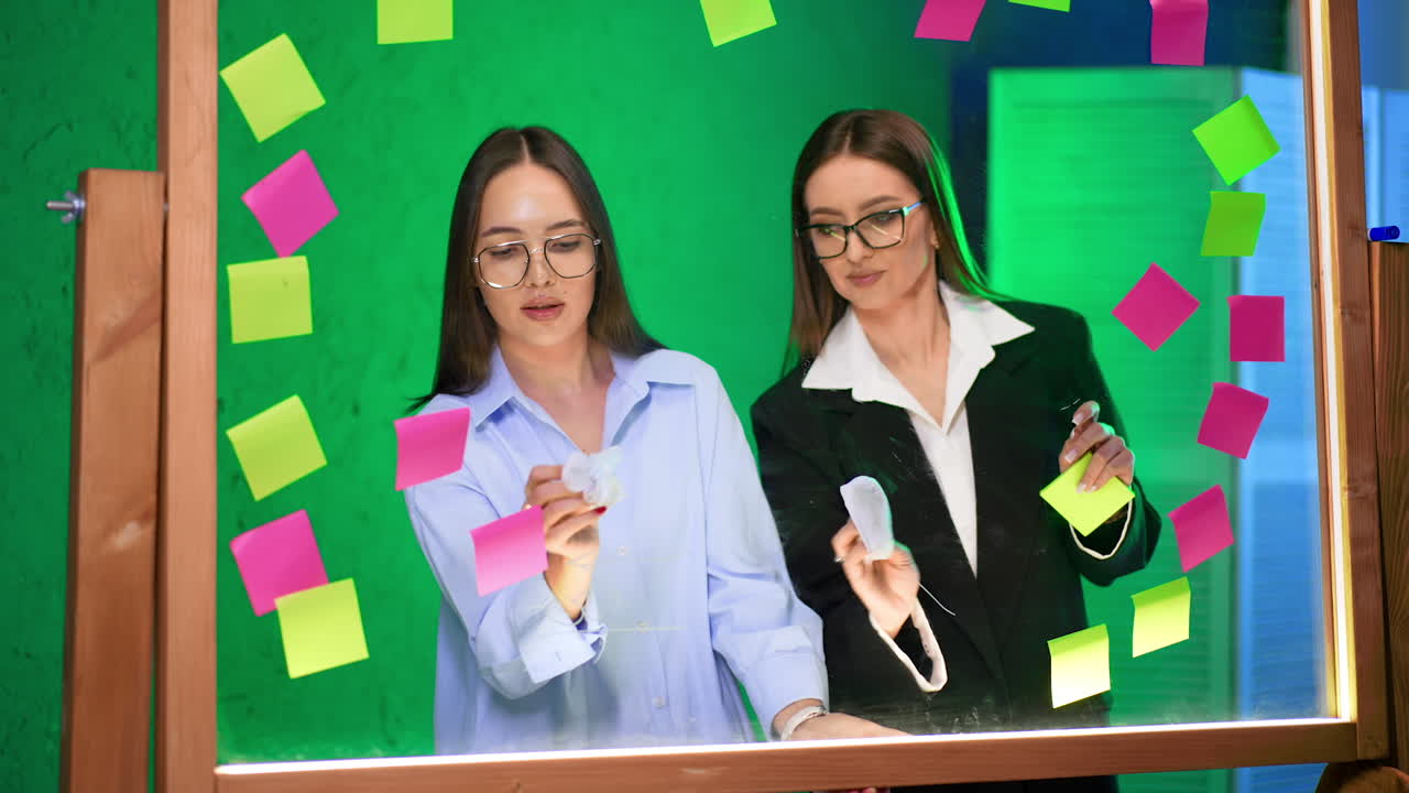 Women brainstorming in studio. Two women collaborate at a glass table, writing on sticky notes surrounded by vibrant colors in a creative space