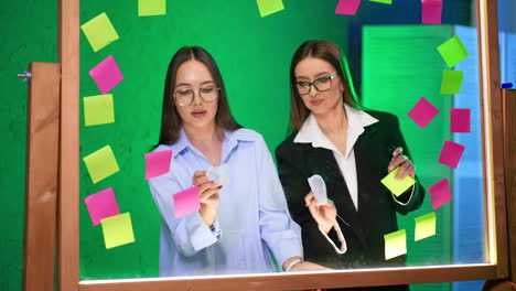 women brainstorming in studio. two women collaborate at a glass table, writing on sticky notes surrounded by vibrant colors in a creative space