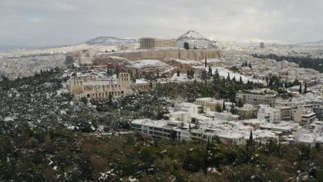 aerial view of the snow covered acropolis hill, blizzard aftermath, in cloudy athens - tracking, drone shot