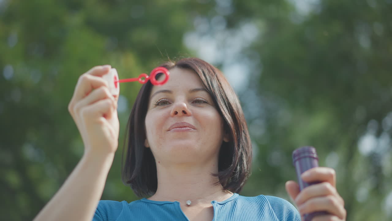 Donna che fa le bolle di sapone in un parco, top blu, primo piano ravvicinato da posizione ribassata, capelli corti, anello per bolle e flacone visibili, luce solare pomeridiana e bokeh verde, respirazione gioiosa ed espressione giocosa suggerite dalle inquadrature
