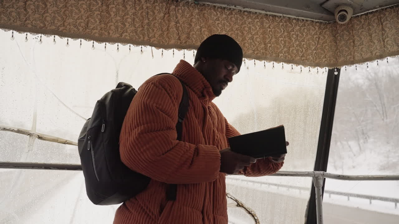 Uomo di colore che legge un libro in un riparo su un balcone innevato, zaino a tracolla, dita che sfogliano le pagine, trama del cappotto di lana, finestra segnata da gocce di pioggia, umore contemplativo, luce soffusa e diffusa