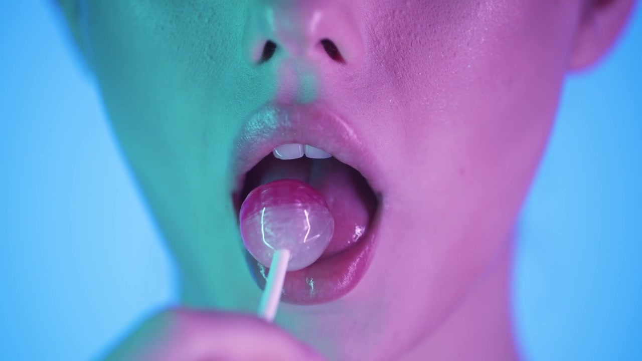 Toma macro 4K en cámara lenta de una mujer lamiendo una piruleta rosa y lamiéndose los labios, fondo azul vibrante con luz de neón rosa y verde brillante, cinemático y juguetón, detalle en primer plano