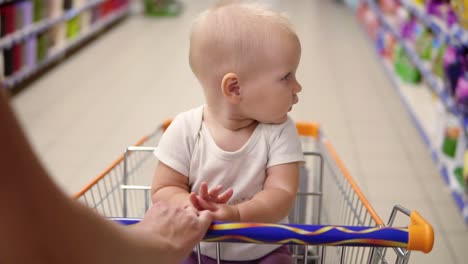 Little-cute-baby-sitting-in-a-grocery-cart-in-a-supermarket-looking-around.-The-mother's-hands-pushing-the-cart-forward-among