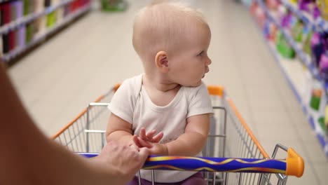 little cute baby sitting in a grocery cart in a supermarket looking around. the mother's hands pushing the cart forward among