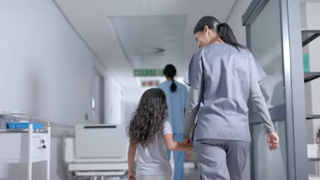 Diverse-female-nurse-and-child-patient-walking-through-corridor-at-hospital,-in-slow-motion