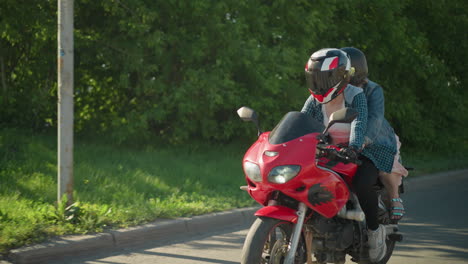 two women ride on a red power bike, the rider's shirt is unbuttoned, showing her white singlet, they pass a signpost while someone walks on the sidewalk, and parked cars