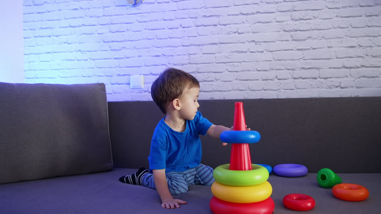 Beautiful Caucasian baby boy sitting on the sofa. Little child playing with a toy pyramid.