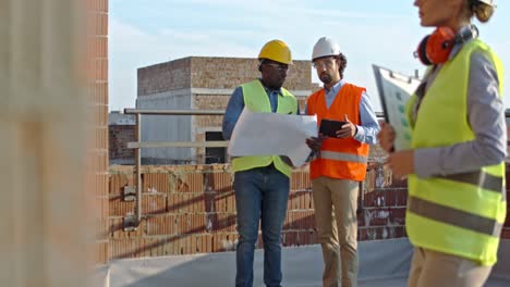 african american and caucasian men constructors in hardhats walking slowly at the building site with drafts of plan and rablet device while talking and discussing next parts of work.