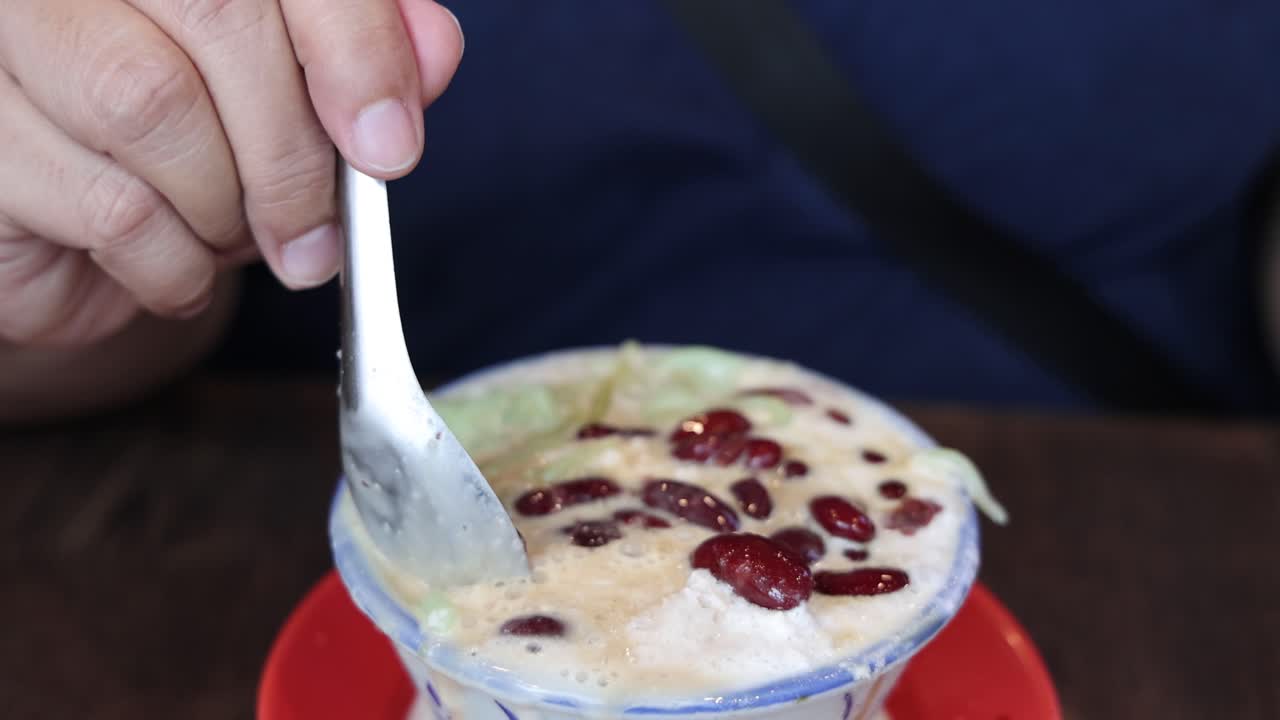 A person uses a spoon to scoop and eat cendol, a shaved ice dessert with red beans, in a brightly lit indoor setting with a close-up view