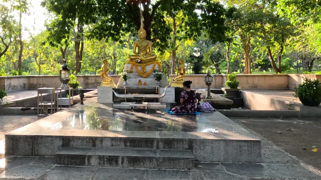 A golden statue surrounded by trees, with sunlight casting dappled shadows on the stone platform.