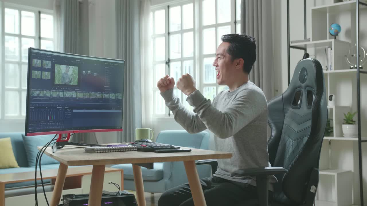 editor de video asiático hombre con camiseta de mangas largas y pantalones negros siendo feliz terminando la edición de video por computadora de escritorio mientras trabaja en casa.