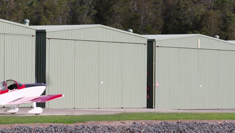 a red and white aircraft is positioned beside a large green hangar, with trees in the background.