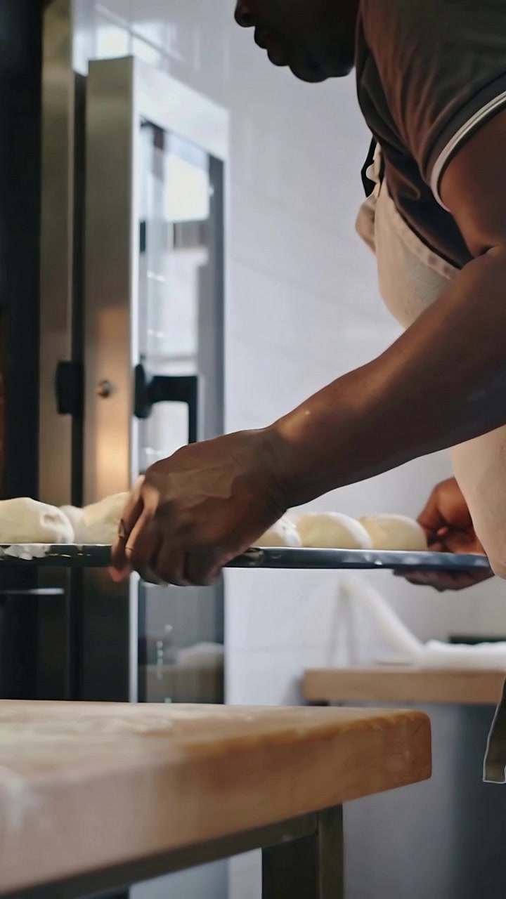 Vertical video: Grasping bake tray, baker placing dough rolls into oven at bakery to bake in apron