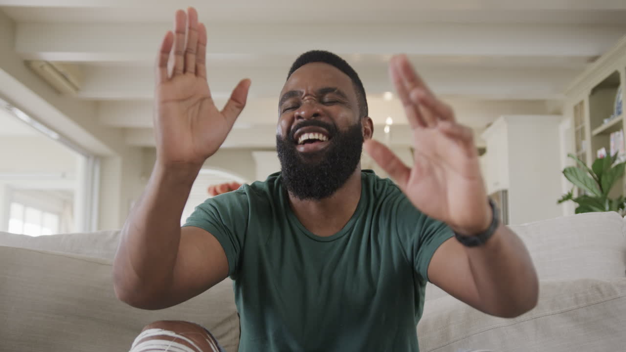 An African American man laughs on a couch in a cozy room on a video call