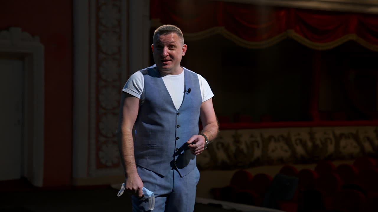 Presenter on stage. Man holding medical mask in his hand and talking on camera in empty theater. Portrait of middle-aged man speaker. Pandemic concept.